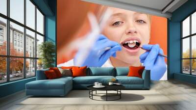 Close-up photo of child girl during dental tooth examination checkup. Attractive pretty caucasian kid sits with opened mouth while cropped female dentist in gloves examining inside of oral cavity Wall mural