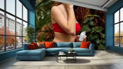 curly female in red bikini applying cream on face in the forest,, enjoys soft skin using organic cosmetics. beauty concept Wall mural