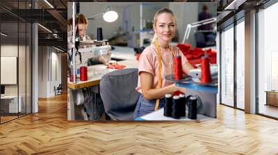 Dressmakers women sewing clothes on sewing machine in factory together, at work. Attractive ladies enjoy tailoring, making clothes, blonde caucasian craftswoman looking at camera smiling Wall mural