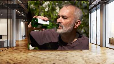 An adult man on a hot day in nature drinks water from a glass bottle. Selective focus. Wall mural