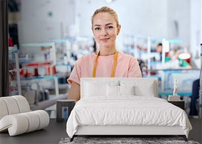 female designer of clothes standing among sewing machines in textile factory, attractive tailor after work stand posing, looking at camera smiling with arms crossed, measuring tape for sewing on neck Wall mural