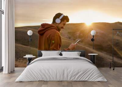 fit caucasian happy man in headphones chatting with someone on smartphone, typing message and smiling outdoors, having rest, enjoy time at sunrise or sunset. in mountains in nature Wall mural