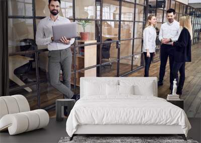 Handsome young man working with laptop in modern office. Hipster in formalwear in coworking. Freelance businessman. A group of people discuss the latest news about the company Wall mural