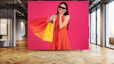 Image of a beautiful excited happy asian woman posing isolated over pink wall background holding shopping bags. dressed in orange dress and sunglasses Wall mural