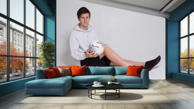 a boy in a white hoodie and black shorts, sitting a white Studio background and holding a ball on his lap Wall mural