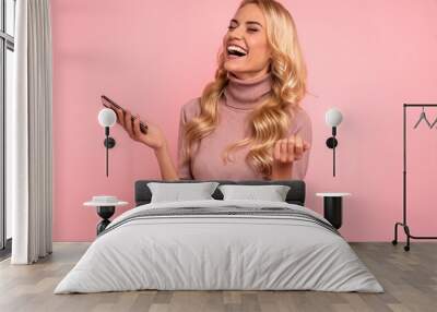 Photo of happy young woman with beautiful long hair laughing and clenching fist while holding mobile phone in hand isolated over pink background Wall mural