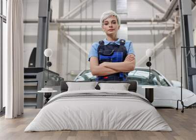 Portrait of adorable caucasian female auto mechanic in uniform posing at camera in auto service, woman with short hair standing with arms folded looking down at camera, alone. Wall mural