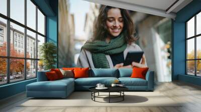 Portrait of young smiling dark-haired woman wearing light-colored coat and green scarf, walking city streets and using smartphone to interact with her friends. Good vibes. Modern technologies concept Wall mural