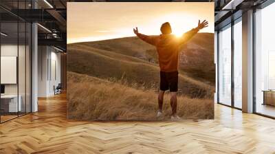 rear view on guy on top of mountain reaches for the sun during sunrise, feel happiness, enjoy the morning outdoors in nature on fresh air. in field. achievement, beginning, happiness, human emotions Wall mural