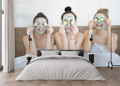 Three funny happy beautiful girlfriends making face clay masks, laughing and eating cucumbers. Stay at home concept. Skin care and treatment, spa, natural beauty and cosmetology concept. Wall mural