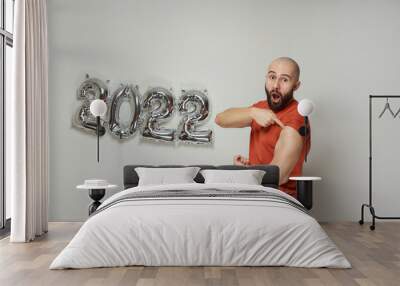 A bald man with a beard and opened mouth is showing with honor a plaster on his shoulder after vaccination against Coronavirus (Covid-19) near foiled silver balloons in the shape of 2022. Wall mural