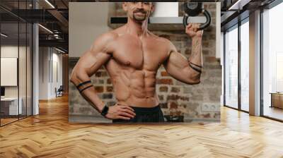 A happy muscular man with a beard is training biceps with a black weight in his apartment. A bodybuilder with a naked torso and tattoos on his arms is warming up during a workout at home Wall mural