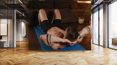 A muscular man with a naked torso is doing an abs workout in his apartment in the evening. An athletic guy with tattoos on his forearms is doing crunches on a blue yoga mat near a couch at home. Wall mural