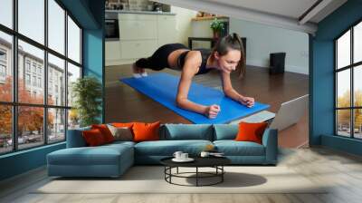 A sporty smiling girl in a black workout tight suit is doing plank watching an online video on a laptop. A female coach conducting a remote fitness class on the blue yoga mat at home. Wall mural