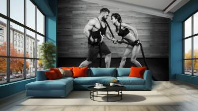 Muscular man with tattoos and beard and attractive girl pulling a rope in a black tank top and black shorts in the gym. Sportive couple doing crossfit exercise. Black and white portrait. Wall mural