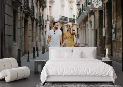 Stylish brunette girl in the hat with her boyfriend with beard walking together in the center of the old European street in Spain in the evening  Wall mural