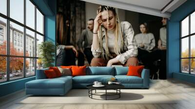 young caucasian alcoholic woman tired of conviction and gossip, sit in the center of room while people in the background discuss problems connected with alcohol. support, problems, help, desperate Wall mural