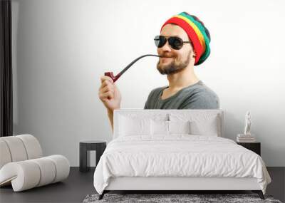 Young caucasian rasta man in jamaica hat, sunglasses and grey t-shirt on white background with smoke pipe in hand. Wall mural