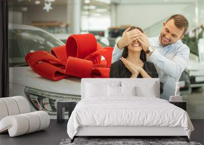 young handsome caucasian guy make surprise to brunette woman, going to give a gift. man closed woman's eyes from back. new car with red bow in the background Wall mural