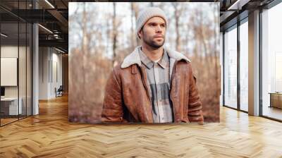 A portrait of a young white man posing in the woods dressed in autumn clothes: brown jacket, checkered canadian style shirt, off-white beanie, looking to the right Wall mural