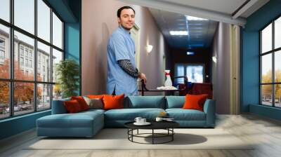 A man who is on the hotel cleaning crew staff is smiling with a towel vacuum in the process of cleaning the hotel rooms and delivering top-knotch service to the guests. Wall mural