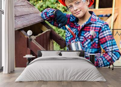 Boy wearing construction helmet with a paint brush ready to apply a varnish on an old wooden dog house. Wall mural