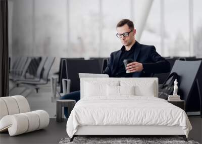 Handsome young businessman working on a laptop and drinking a coffee during an expectation of a flight Wall mural