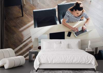 Top view of an adult woman sitting at desk and working on her laptop, her table is perfectly tidy, the sun casts graphics shadows on the wood floor. Wall mural