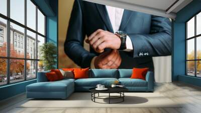 A close-up of a cropped frame of a man in an expensive classic costume looks at his watch. The businessman is in a hurry and looks at the time. A handsome man with a precious watch and a gold ring. Wall mural