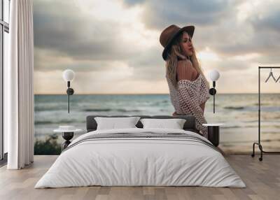 Rear view of a sexy girl in a brown swimsuit and a hat on her head posing on the beach at sunset, shows her beautiful ass. A young model walks and sunbathes near the ocean on the sand. Travel concept. Wall mural