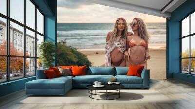 Two beautiful blonde girls with sunglasses on the face, sexy bodies are resting on a windy beach at sunset. Women in swimsuits on an island near the ocean. Travel concept. Wall mural