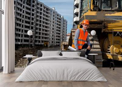 a great worker in uniform and helmet stands by an excavator on a construction site. Wall mural