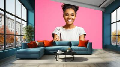 Smiling girl holds hands crossed. Photo of african american girl wears casual outfit on pink background. Emotions and pleasant feelings concept. Wall mural