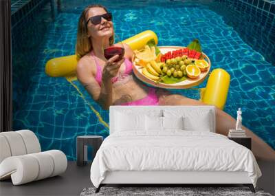 Beautiful woman lying on floating hammock in the swimming pool with wine and tray of fruits Wall mural