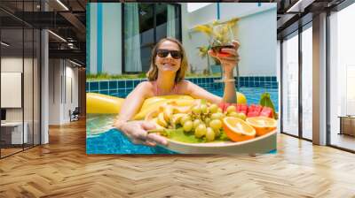 Beautiful woman lying on floating hammock in the swimming pool with wine and tray of fruits Wall mural