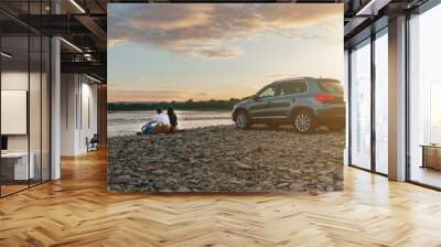 Portrait of happy young adult couple with dog on roadtrip. Man sitting on plaid with woman and watching sunset near river. Outdoor picnic concept. Wall mural