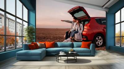 Young Happy Couple Dressed Alike in White Shirt and Jeans Sitting at Their New Car Trunk, Beautiful Sunset on the Field, Vacation and Travel Concept Wall mural