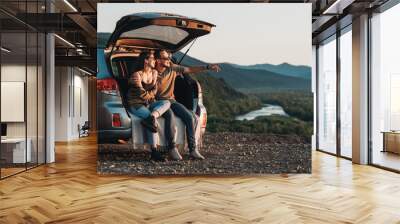 Young Traveler Couple on a Road Trip, Man and Woman Sitting on the Opened Trunk of Their Car Over Sunset Wall mural