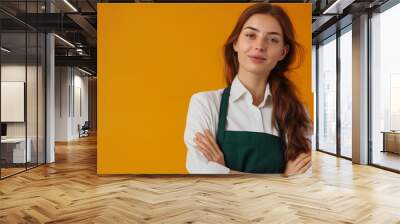  beautiful young woman in white shirt and green apron with arms crossed looking at camera over yellow background, copy space concept, barista uniform service industry  Wall mural