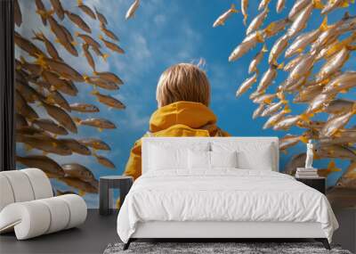 A child in a yellow jacket stands captivated by a school of fish swimming against a bright blue sky. Wall mural