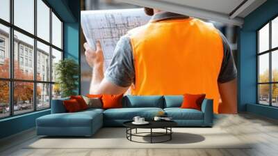 A construction worker in an orange safety vest and hard hat inspects blueprints at a building site. Wall mural