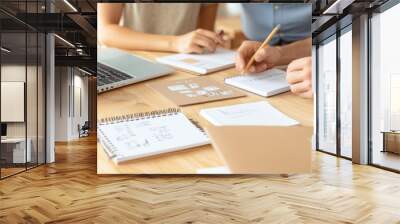 Collaborative workspace scene with hands writing notes, using a laptop, and organizing creative ideas on a wooden desk. Wall mural