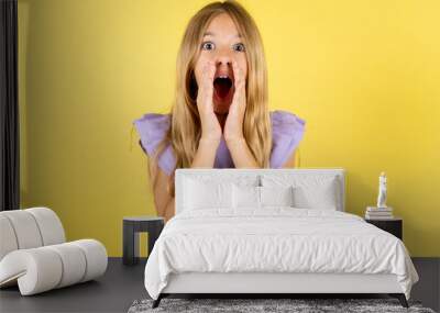 blonde kid girl wearing violet T-shirt over yellow studio background shouting excited to front. Wall mural