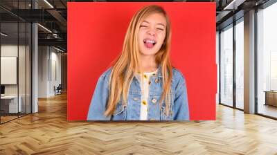 blonde little kid girl wearing denim jacket over red background sticking tongue out happy with funny expression. Emotion concept. Wall mural