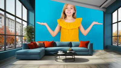 Cheerful cheery optimistic Caucasian kid girl wearing yellow T-shirt against blue wall holding two palms copy space Wall mural