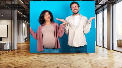 Cheerful cheery optimistic young couple expecting a baby standing against blue background holding two palms copy space Wall mural