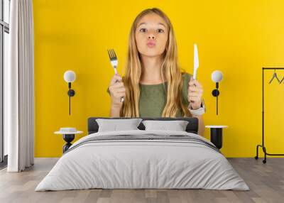 hungry blonde kid girl wearing green T-shirt over yellow studio background holding in hand fork knife want tasty yummy pizza pie Wall mural