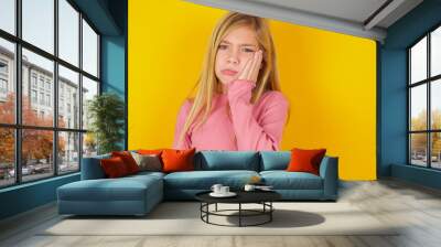 Sad lonely caucasian little kid girl wearing long sleeve shirt over yellow background touches cheek with hand bites lower lip and gazes with displeasure. Bad emotions Wall mural