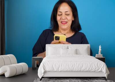 Smiling middle aged Arab woman standing against blue background using cell phone, messaging, being happy to text with friends, looking at smartphone. Modern technologies and communication. Wall mural