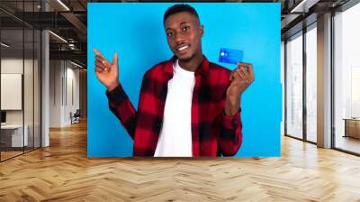 Smiling young handsome man wearing overshirt over blue background showing debit card pointing finger empty space Wall mural